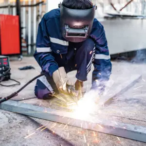 A welder wearing protective gear and a dark welding helmet performs hot work on a metal plate in an industrial outdoor setting, with sparks flying from the welding torch and welding equipment visible in the background.
