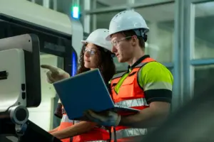 Two workers in hard hats and safety vests reviewing a laptop near industrial machinery