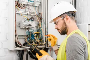 Electrician in hard hat and safety vest testing wires in an electrical panel with a multimeter