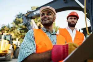 Two construction workers in hard hats and orange vests standing near heavy equipment on a job site
